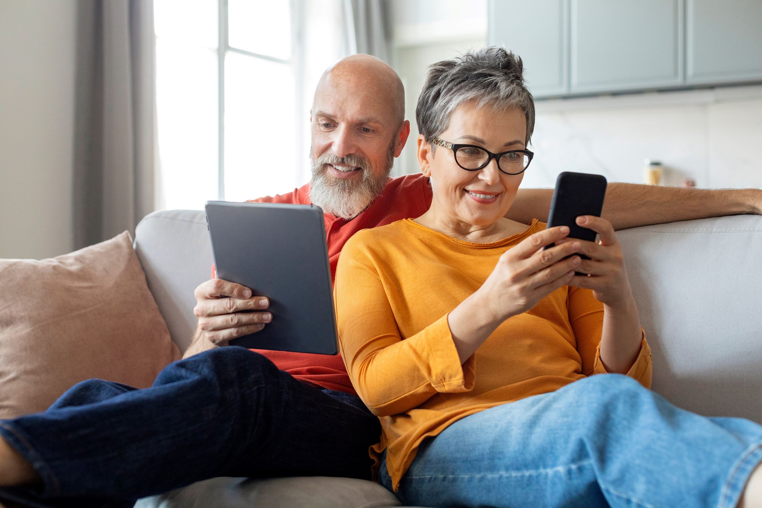 Senior citizen man and woman sitting on couch reading a tablet and cell phone
