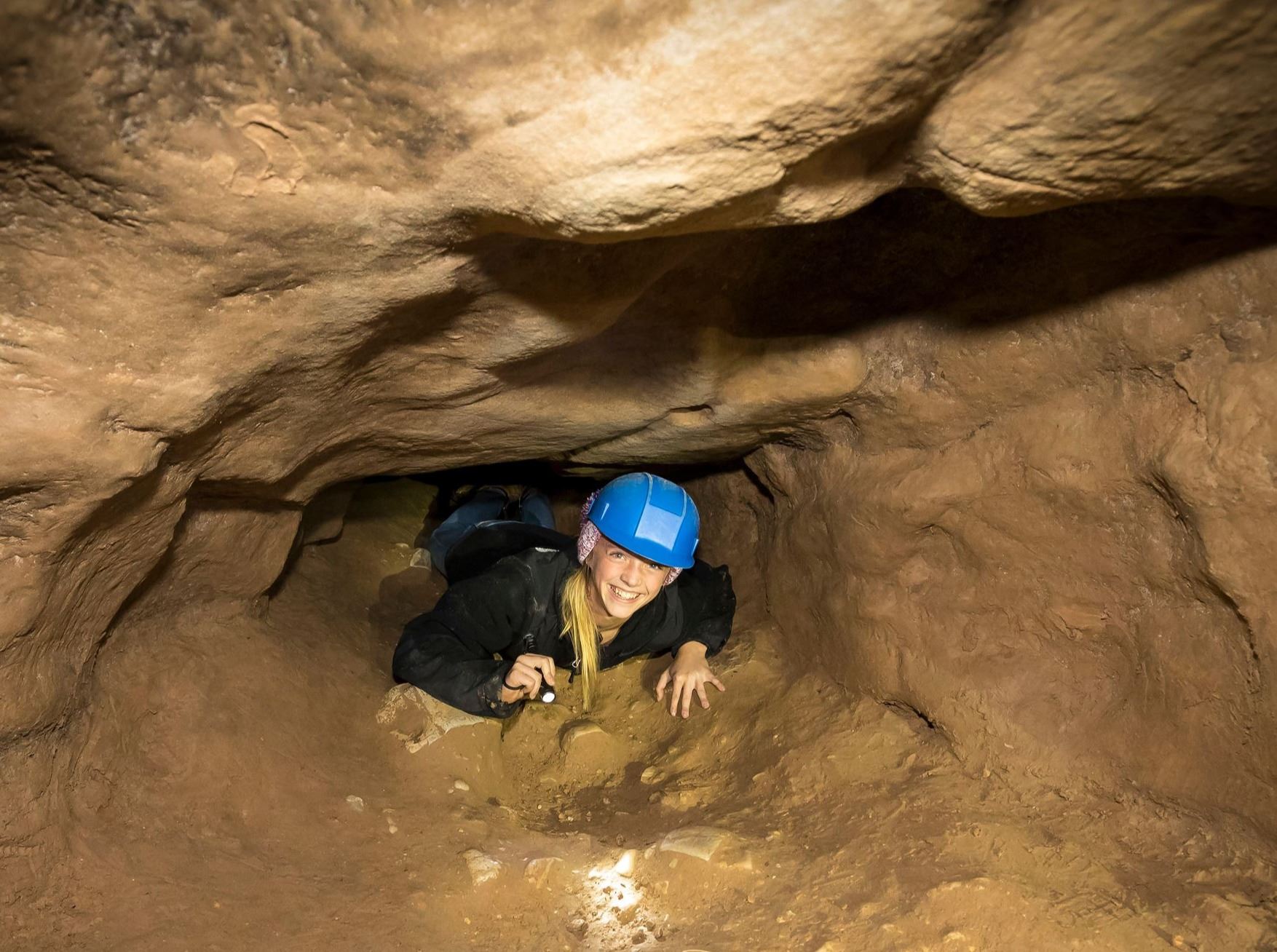 A girl wearing a blue helmet and holding a flashlight is crawling on her belly through a tunnel in a